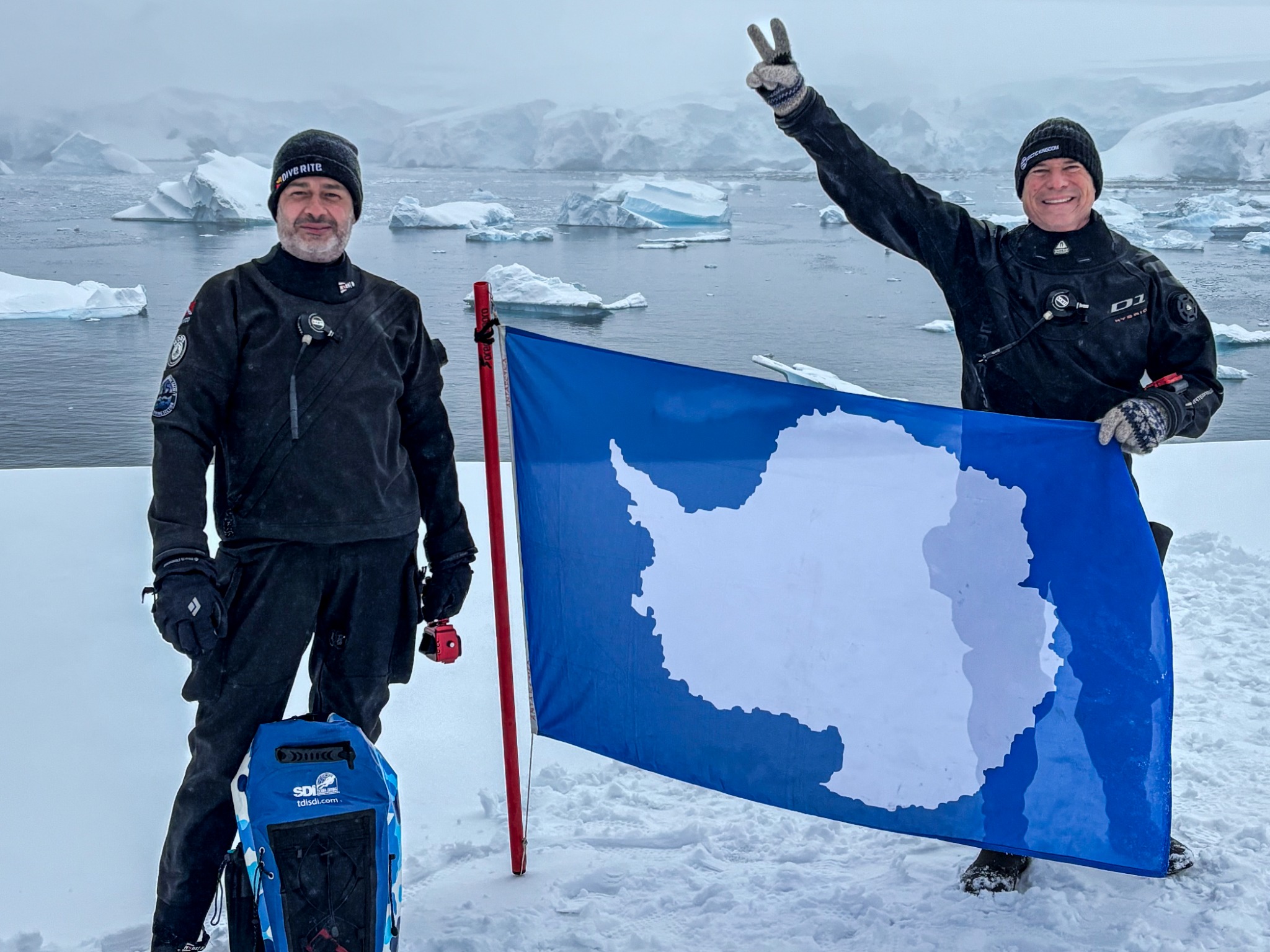 Two polar divers standing on Antarctic ice with an Antarctica flag